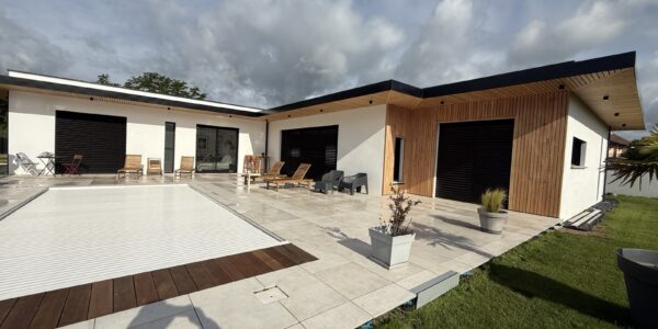 Modern single-story house with white walls and wood cladding, a wide tiled patio, and a retractable pool cover extending to a grassy lawn.