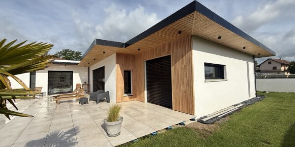 Modern single-story house with white walls, vertical wood cladding, and a large overhanging roof; a tiled patio with outdoor seating and planters.