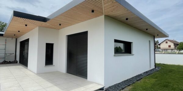 Modern white house exterior with flat roof, wood soffit, and large black roller shutters over windows and doors; tiled patio adjacent to green lawn.