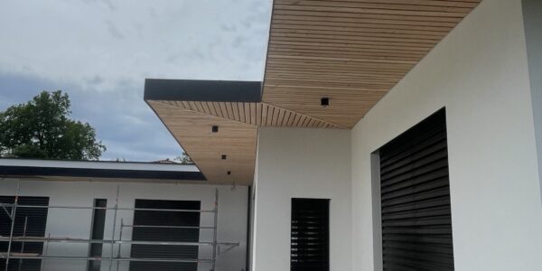 Modern building exterior with long wooden eaves, white walls, and large tiled courtyard; scaffold visible in background indicating ongoing construction/finish work