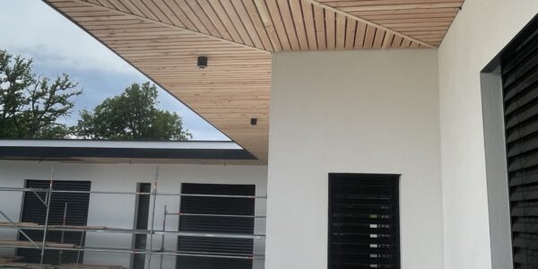 Wood-paneled overhang on a modern building with white walls and black louvered windows under a bright sky.