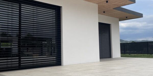 Modern house facade with white walls, black-framed glass doors, and a wooden slatted overhang above a large tiled patio.