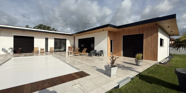 Modern single-story house with wooden cladding on a patio, lounge chairs, and a covered pool in a sunny yard.
